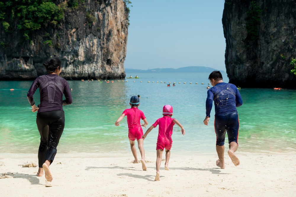 Kids playing at a Phuket beach with rashguards and sun hats, accompanied by their parents.
